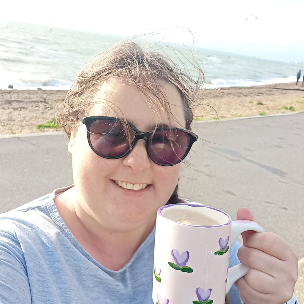 Elizabeth stands on a beach smiling and holding a cup of tea